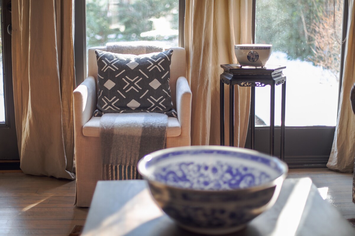 A softly lit corner features a cozy armchair draped with a striped blanket and adorned with a patterned throw pillow. In front, a wooden table holds a decorative bowl, while sunlight illuminates the space, highlighting the outdoor view through large windows.