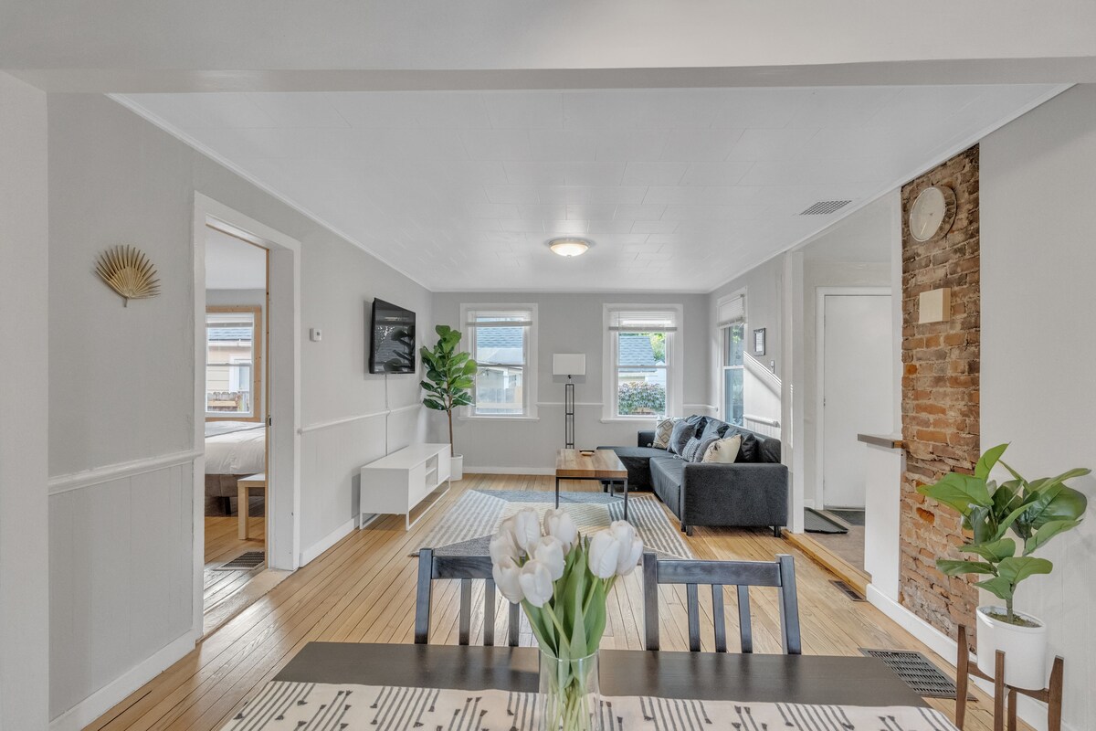 A spacious living area features large windows allowing natural light to illuminate the neutral-toned walls and hardwood flooring. A comfortable sofa is positioned beside a modern television unit, and a dining table adorned with fresh flowers offers a warm touch.