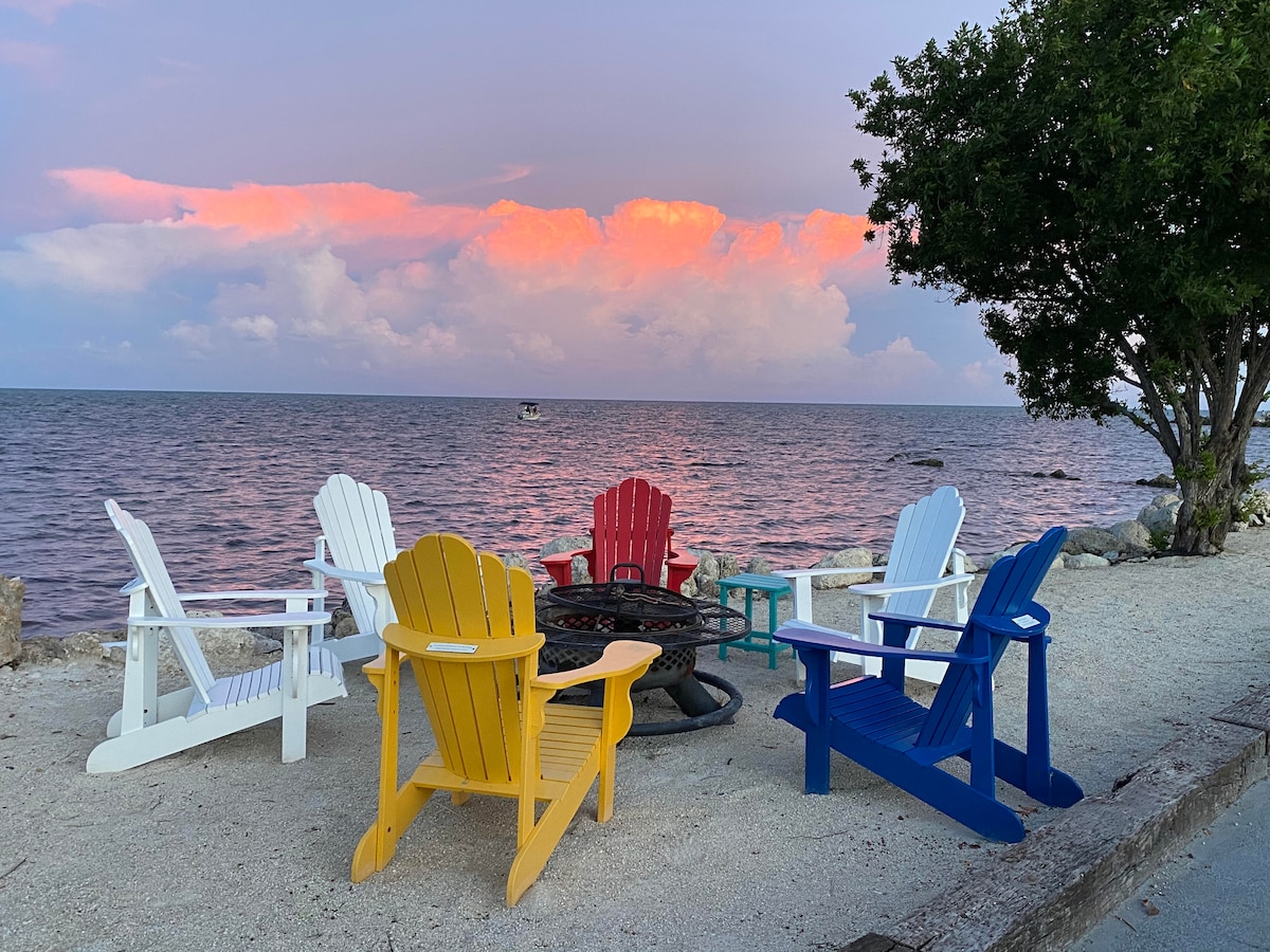A circle of brightly colored Adirondack chairs in red, yellow, blue, and white surrounds a fire pit, facing the tranquil waters. The calm sea reflects soft pastel hues of the sky at sunset, while fluffy clouds are visible in the background.