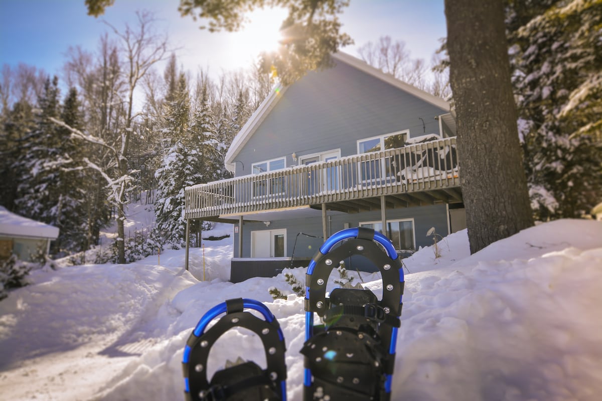A cozy chalet is nestled among snow-covered trees, featuring a spacious balcony that overlooks a serene winter landscape. Snowshoes rest in the foreground, indicating access to winter activities. The sun shines brightly above, casting a warm glow on the exterior of the home.