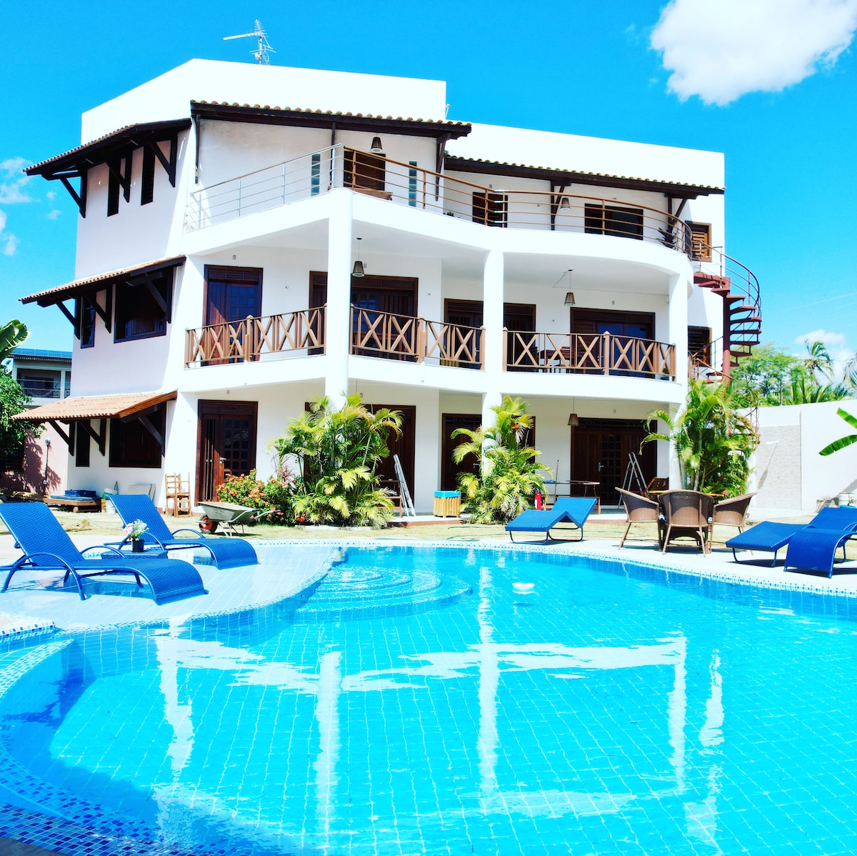A multi-story guesthouse is depicted overlooking a clear blue swimming pool. The outdoor area includes sun loungers arranged around the pool, while lush greenery adds a natural touch. The building features a combination of balconies and outdoor seating areas, complemented by bright blue skies.