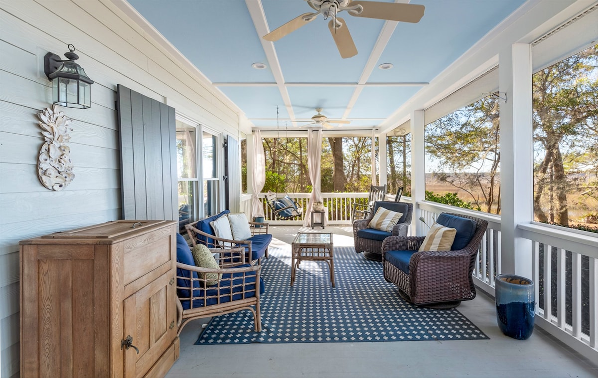 A spacious screened-in porch features comfortable seating with blue and neutral cushions arranged on a patterned rug. A wooden coffee table sits in the center, while ceiling fans and large windows provide ventilation and views of the surrounding greenery.