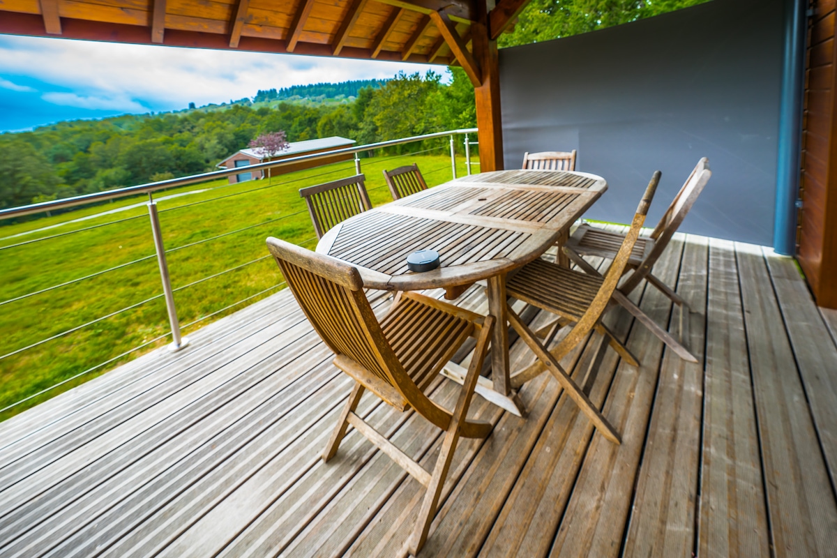A wooden terrace features a large dining table surrounded by six chairs, overlooking a lush green landscape. The area is sheltered by a wooden roof, with a smooth wooden floor enhancing the outdoor setting. The scenery includes gentle hills in the background.