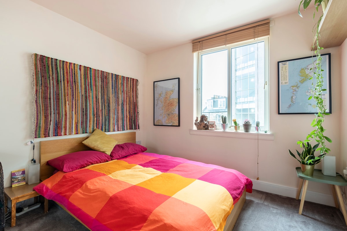 A brightly coloured bedspread covers a spacious bed, complemented by decorative pillows. Natural light fills the room through a large window, which is dressed with light blinds. Plants are positioned on the windowsill, while framed maps hang on the walls.