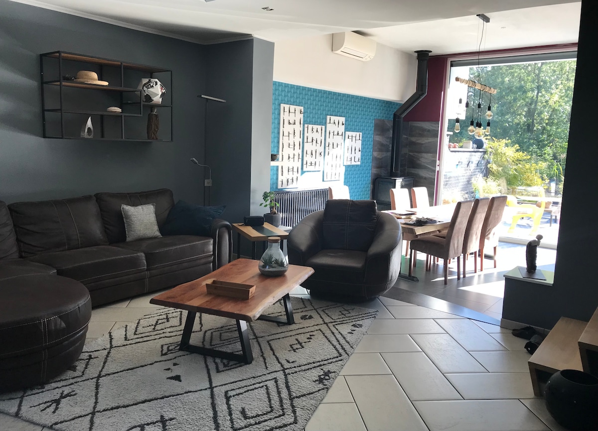 An open living area combines a modern lounge with a dining space. A comfortable seating arrangement features a sectional sofa and armchairs arranged around a wooden coffee table. Large windows provide views of the garden, with blue accent walls enhancing the decor.