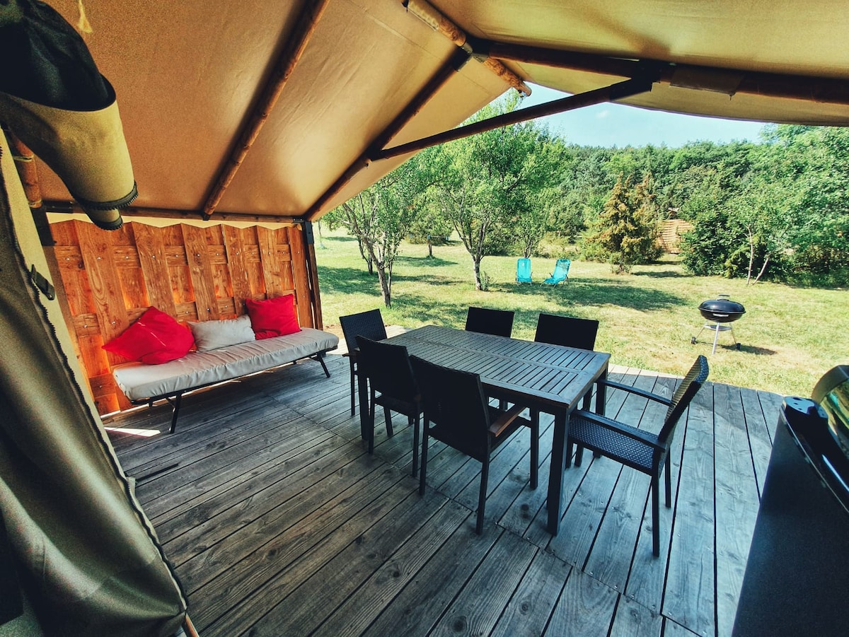 An outdoor terrace features a wooden dining table surrounded by black chairs, with a lounge area showing a cushioned sofa. Soft red pillows enhance the seating. A portable grill stands nearby, and a grassy area with trees is visible beyond the deck.