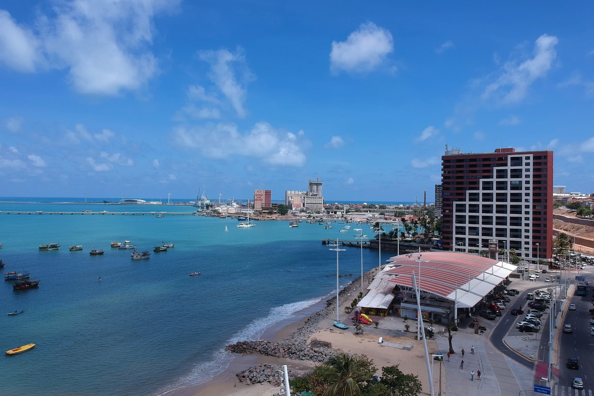 The image captures a scenic coastal view displaying clear blue waters and a vibrant shoreline. Several boats are anchored in the harbor, while the skyline features a mix of modern buildings and historic structures, creating an inviting atmosphere by the beach.