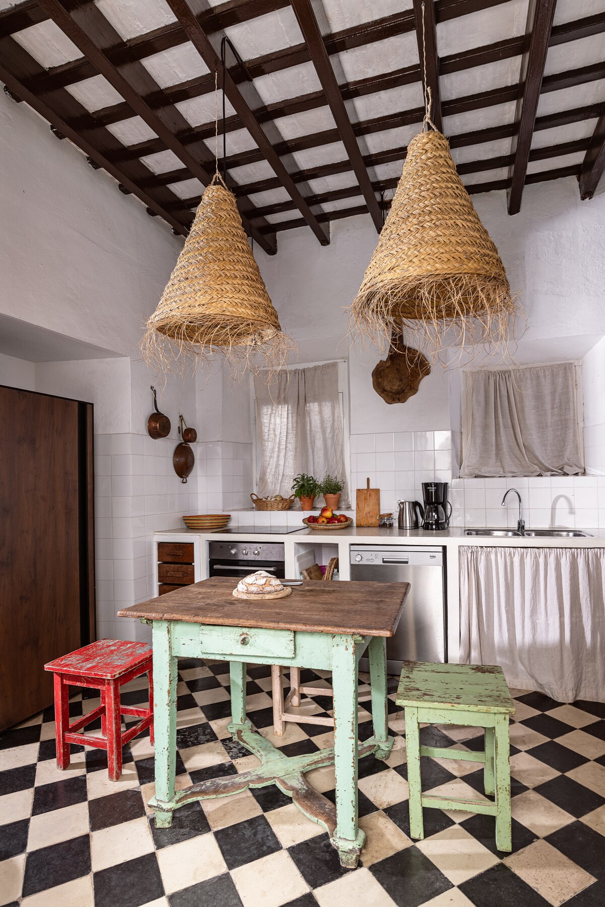 The kitchen is designed with a contrasting black and white checkered tile floor. Two large woven pendant lights hang from the beamed ceiling. A rustic wooden table is surrounded by colorful chairs, while modern appliances are integrated into the cabinetry.