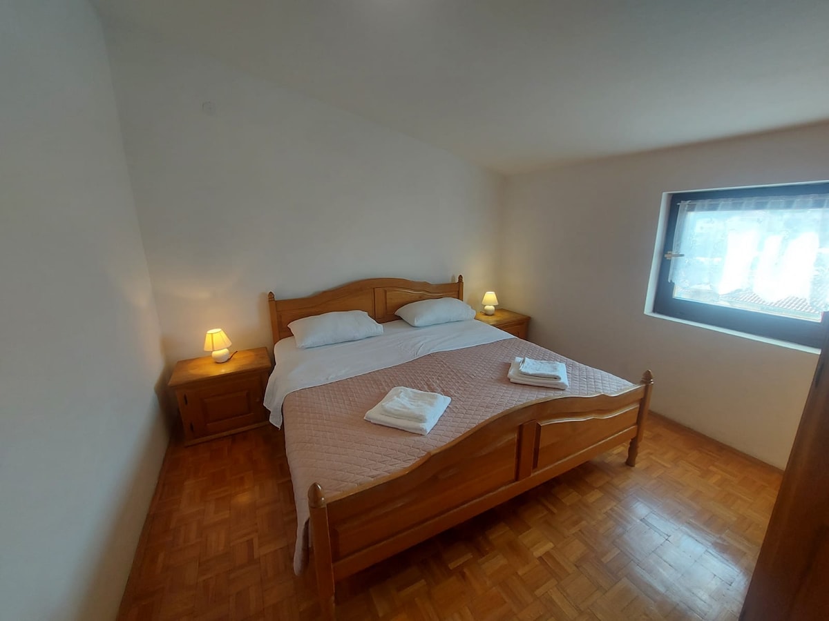 A bright bedroom features a spacious bed with light linens and two bedside tables, each with a lamp. Natural light enters through a window with sheer curtains, illuminating the wooden flooring and simple decor.
