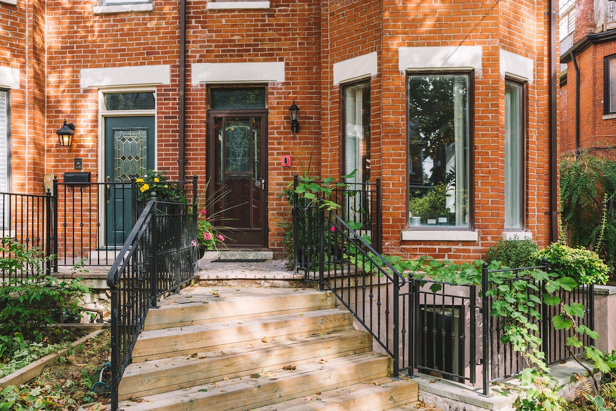 The exterior of the sun-filled unit features a red brick facade with large windows flanking the entrance. A set of wooden stairs leads up to a central door, framed by decorative elements. Lush greenery and flowering plants are positioned along the pathway and railings.