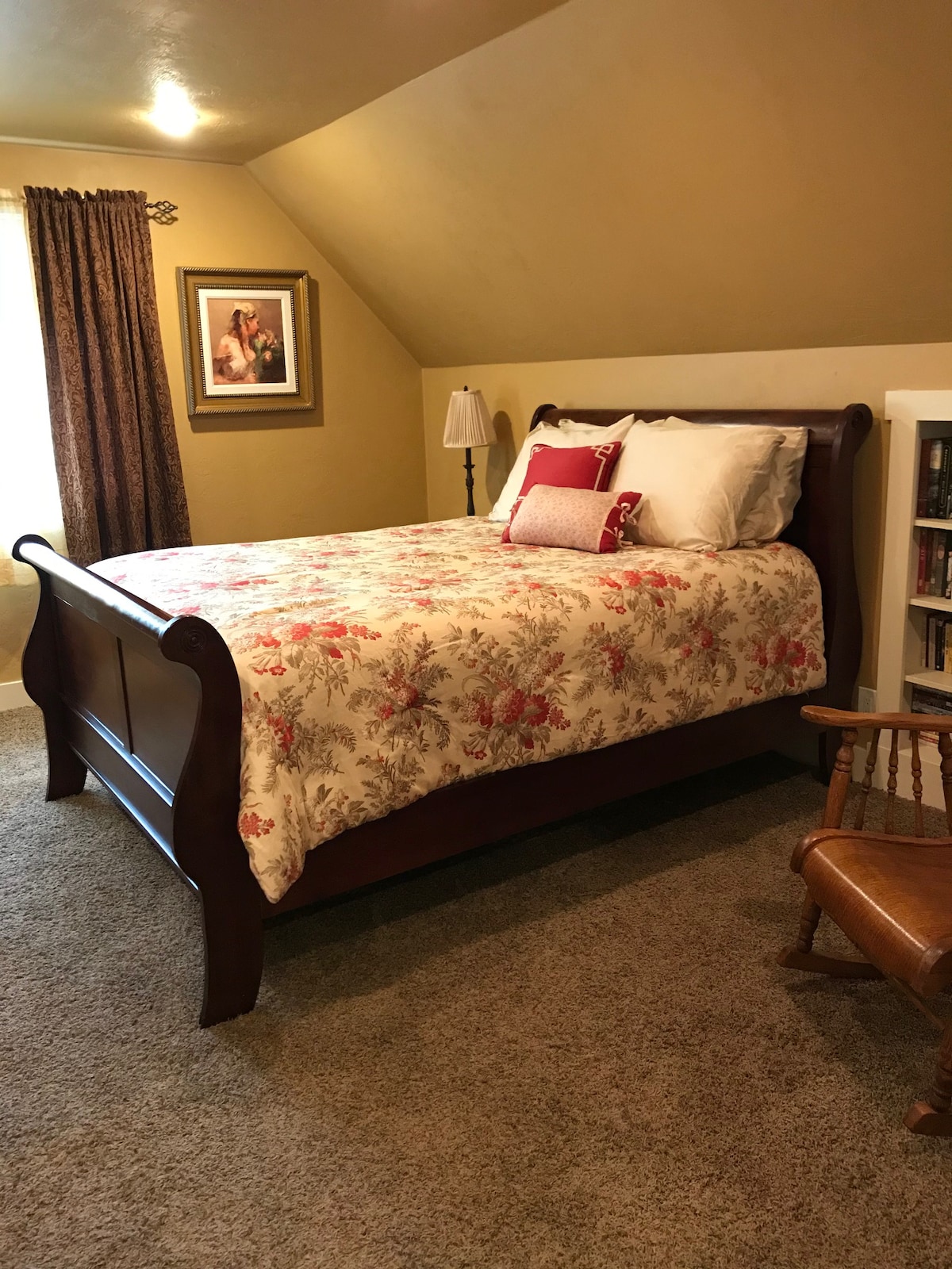 A cozy bedroom features a wooden sleigh bed with a floral-patterned quilt and decorative pillows. A small bedside lamp and a rocking chair are positioned nearby. Soft light illuminates the room, and a framed artwork adds a touch of elegance to the wall.