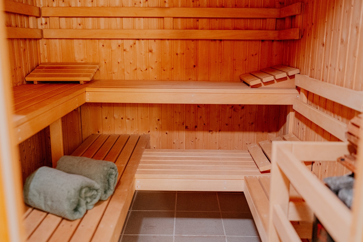 The sauna space features wooden benches arranged for relaxation, accompanied by two folded towels. Warm wooden panels surround the interior, while the floor is finished with tiles, providing a serene environment for unwinding.