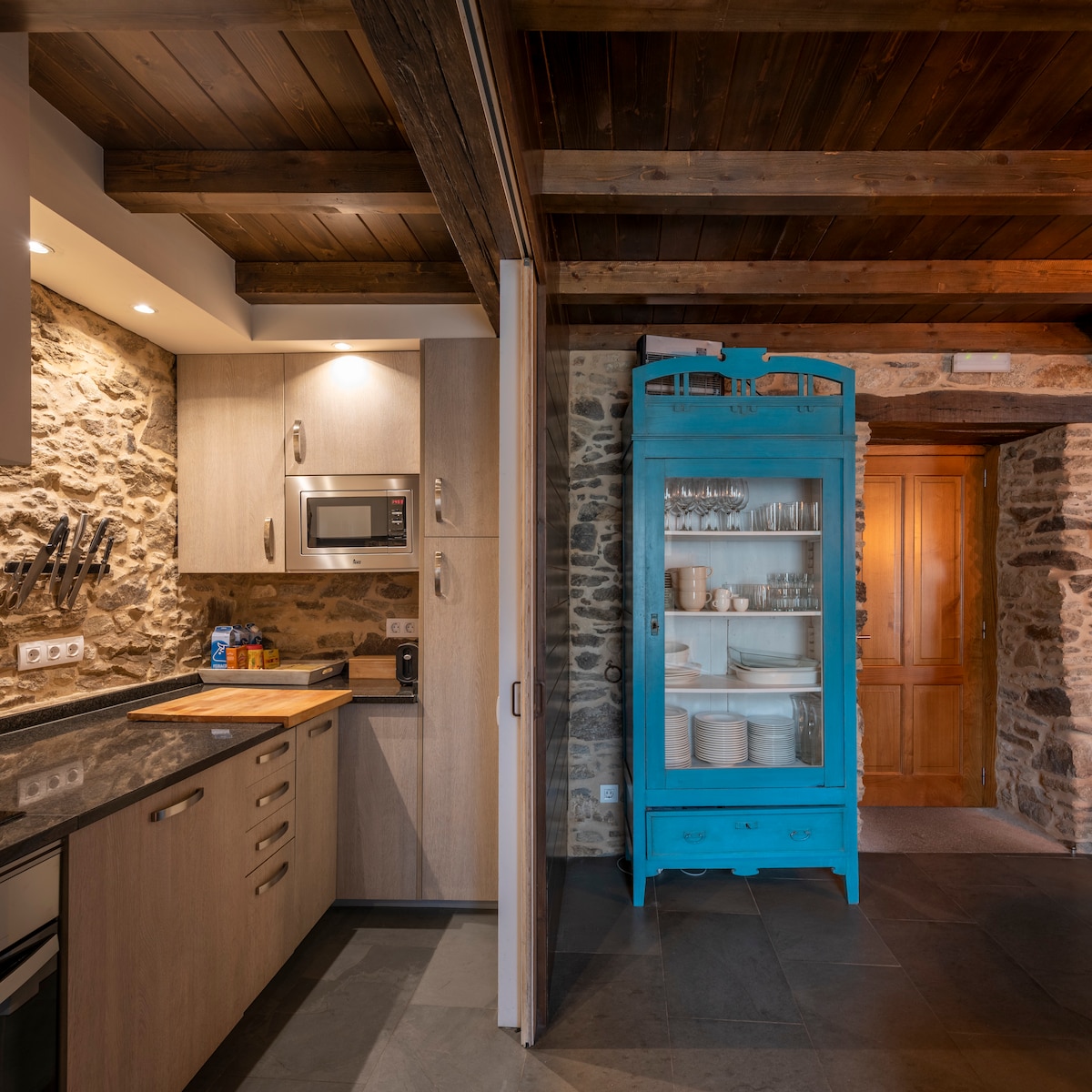 The kitchen area features stone walls and wooden beams, creating a rustic atmosphere. Equipped with modern appliances, including a microwave and cooking area, the design includes a vibrant blue cabinet displaying various dishes and glassware, enhancing the space's charm.