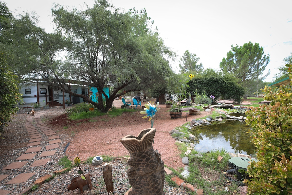 A serene outdoor garden space features a winding stone path leading to a tranquil pond surrounded by greenery. Sculptures and decorative elements are placed throughout the area, with seating arrangements set beneath the shade of trees, promoting relaxation and connection with nature.