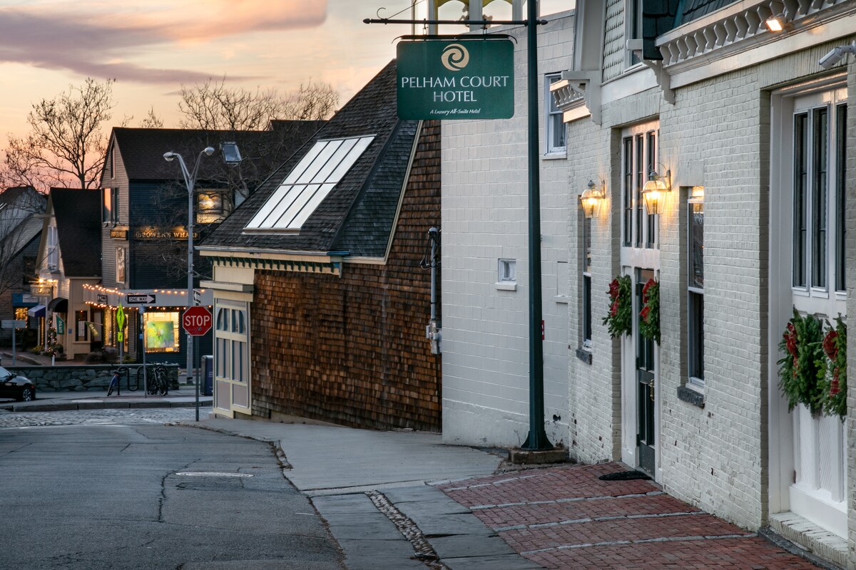 The image captures the exterior of Pelham Court Hotel at dusk, with soft lighting illuminating its entrance. Seasonal wreaths are displayed on the building, and a gentle street incline is visible, leading towards shops in the background. A stop sign is seen along the street, enhancing the urban atmosphere.