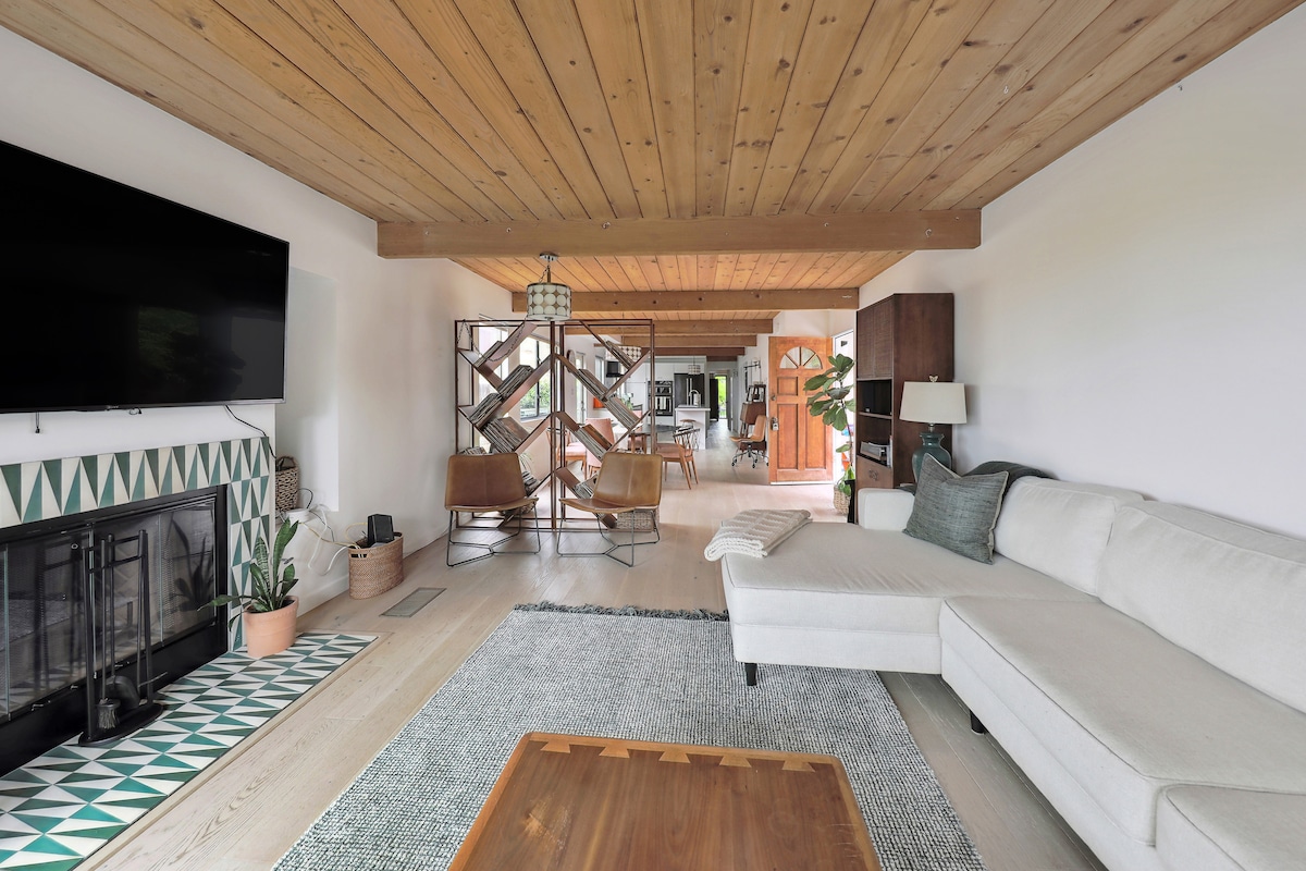 A spacious living area is highlighted by a cozy sectional sofa and a wooden coffee table. Natural light floods through large windows, complementing warm wooden beams on the ceiling. A patterned tile fireplace adds a touch of elegance, while greenery adds freshness to the space.