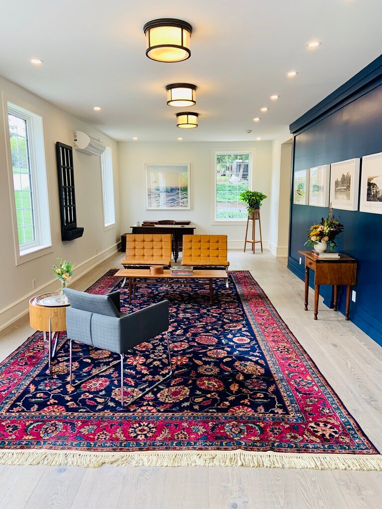 A modern lounge space features a rich, patterned rug and elegant seating arrangements. Mid-century style chairs are complemented by a sleek table. Natural light is provided by large windows, enhancing the welcoming atmosphere of the room, adorned with framed artwork and decorative plants.
