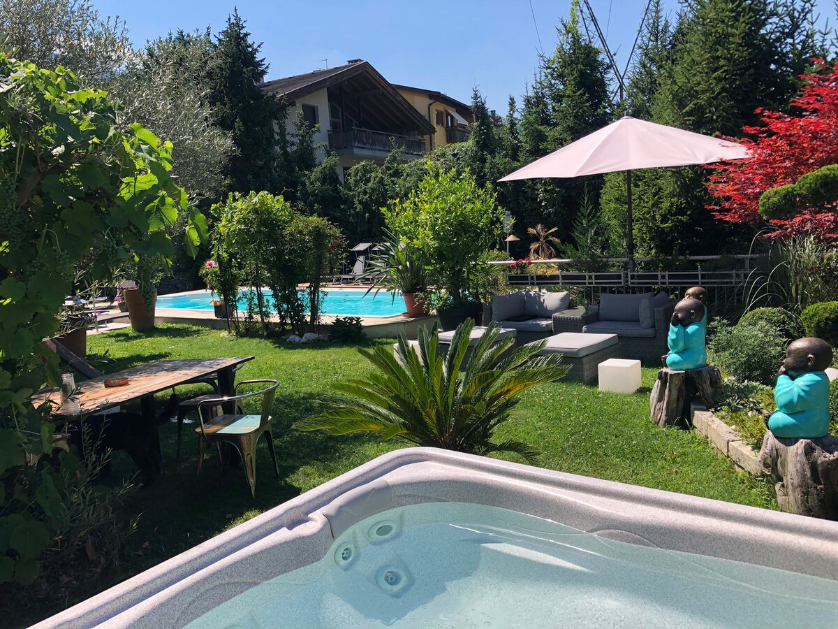 A lush garden oasis is showcased, featuring a large swimming pool surrounded by greenery. Comfortable seating areas with modern furnishings are visible, and a hot tub is positioned nearby under a shaded umbrella. Decorative plants and sculptures add a touch of tranquility to the outdoor space.