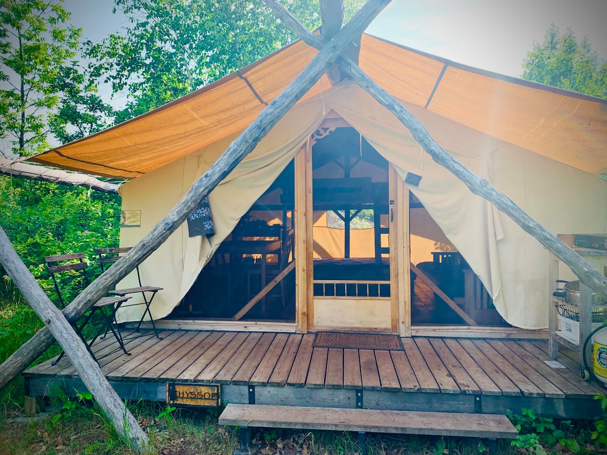 A canvas cabin features a wooden deck with a small chair set outside. The entrance is framed by large wooden beams, while light filters through the open flaps, revealing a spacious interior. Nature surrounds the cabin, enhancing the rustic appeal.