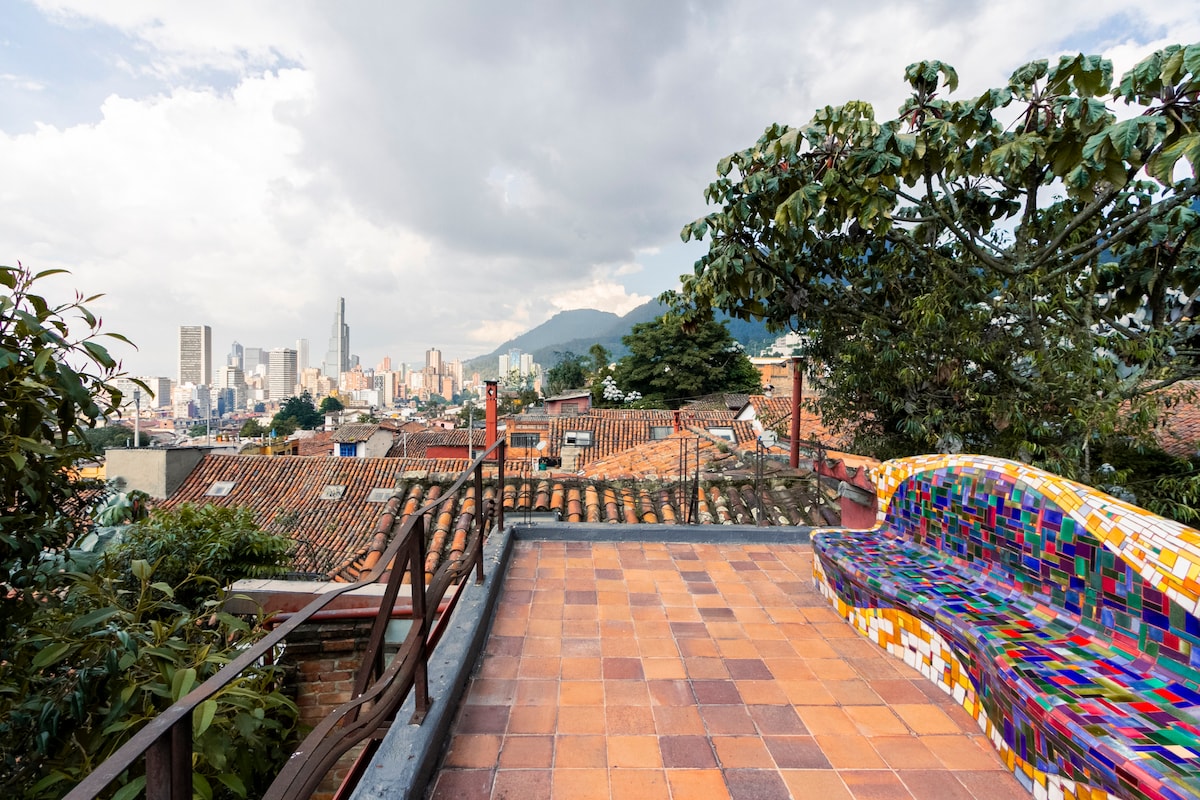 A vibrant rooftop terrace features a colorful mosaic bench overlooking the cityscape. A blend of traditional and modern architecture is visible in the distance, framed by lush greenery and mountains. The textured patio adds warmth to the inviting space.
