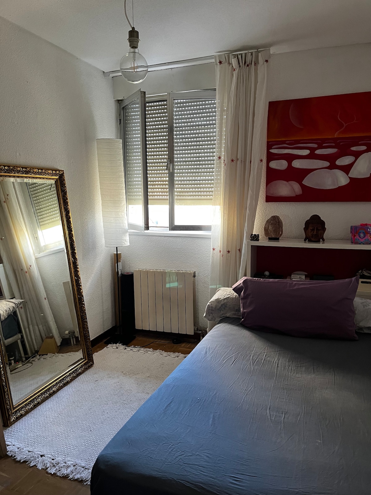 The room features a spacious bed with a purple accent pillow, complemented by a large mirror reflecting the ambient light. Natural light enters through the window, adorned with sleek blinds, while a warm area rug rests on the wooden floor beside the bed.
