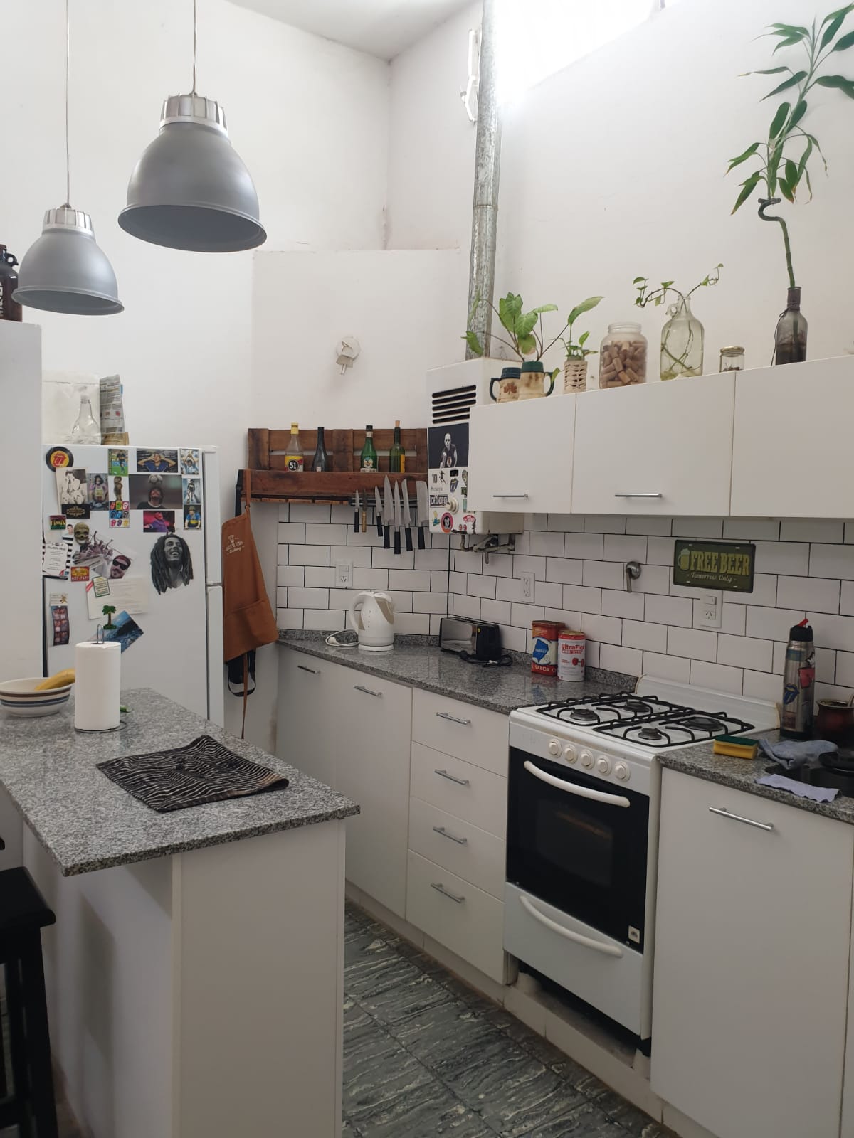 The kitchen features a modern design with white cabinetry and gray countertops. Natural light is provided by a window, highlighting a cooking space equipped with a stove, sink, and various utensils. Decorative items, including plants and wall accessories, add a personalized touch.