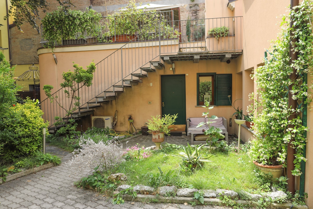 A private garden area features a mix of potted plants and greenery. An outdoor seating space is included, accompanied by two staircases leading to an upper level. The entrance door is framed by green shutters, contributing to the serene atmosphere of the courtyard.