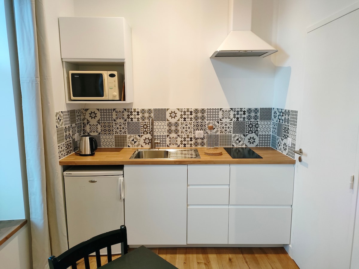 A compact kitchen area features a mix of modern appliances including a microwave and kettle. The tiled backsplash displays a unique pattern, while a wooden countertop provides space for meal preparation. Storage cabinets are positioned above and below the counter for convenience.