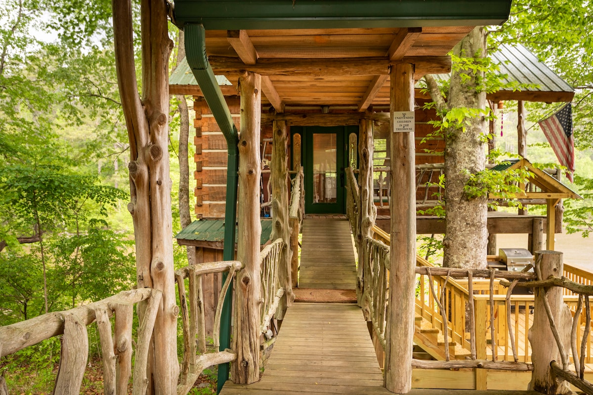 A charming wooden walk bridge leads to the entrance of the tree cabin, surrounded by lush greenery. The structure integrates natural elements, with tree trunks and branches artistically incorporated into the design, inviting guests to explore the tranquil environment.