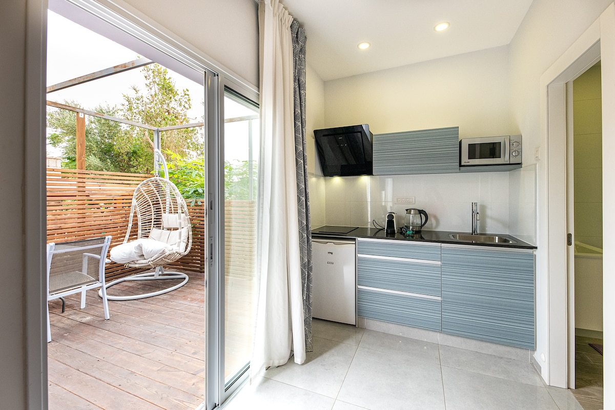 A modern kitchen is presented with sleek cabinets and integrated appliances, including a microwave and refrigerator. A large window allows natural light to fill the space, with a view of a wooden terrace featuring a hanging chair and outdoor seating.