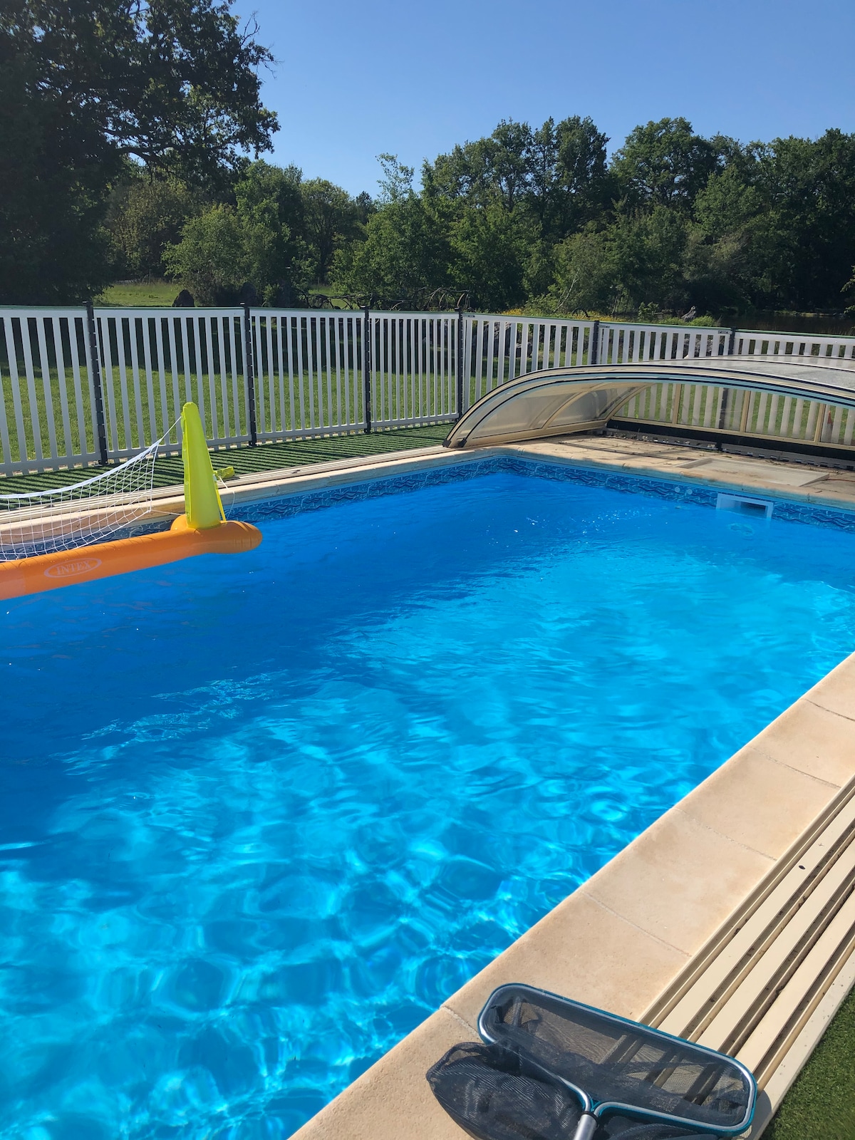 A clear, blue swimming pool is surrounded by a white safety fence. Sunlight reflects off the water, creating a sparkling effect. A protective cover is partially opened, while a bright inflatable pool float rests on the water's surface.