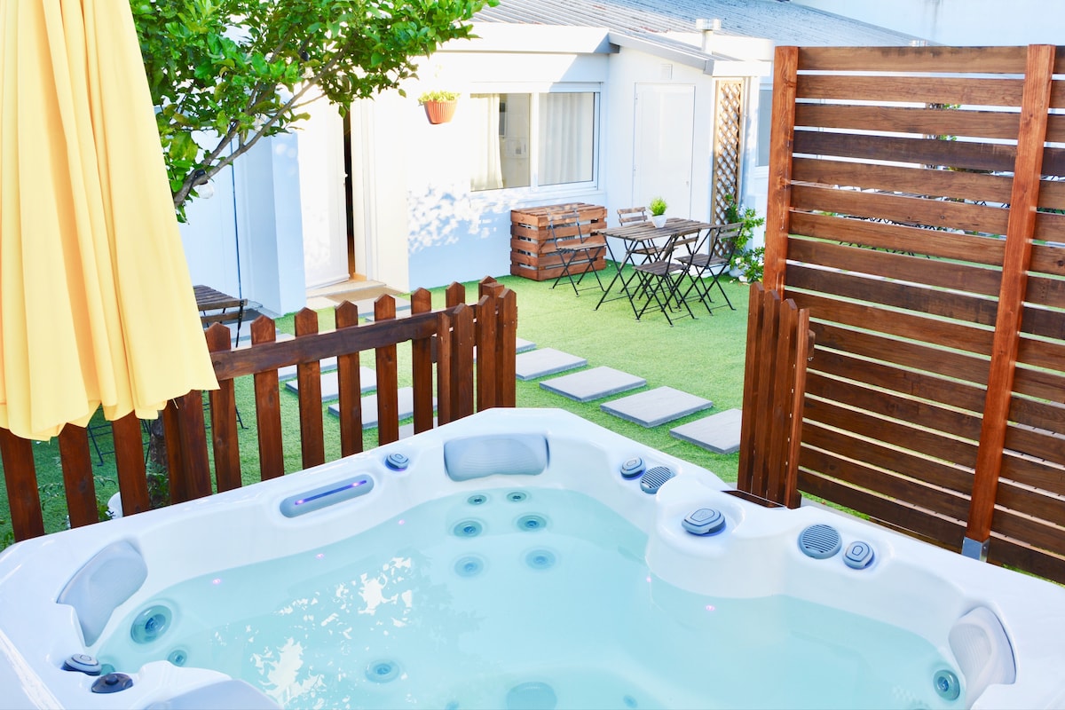 A shared jacuzzi is surrounded by a wooden fence and a sun umbrella. The garden features a stone pathway leading to a seating area with two chairs and a table, enclosed by green grass and a tree offering shade.