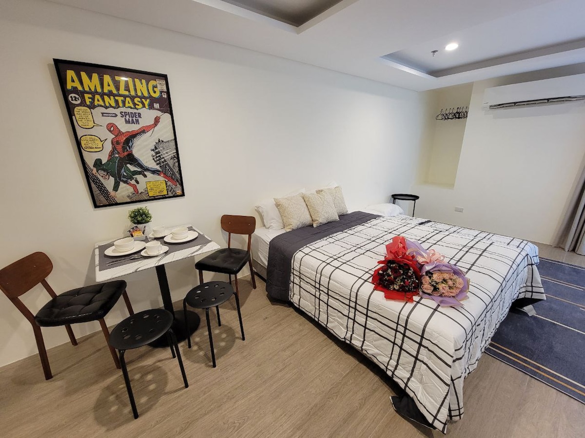 A cozy sleeping area features a spacious bed adorned with decorative pillows. A small dining table set for four is positioned nearby. The wall showcases a vibrant comic book poster, adding a playful element to the modern design.