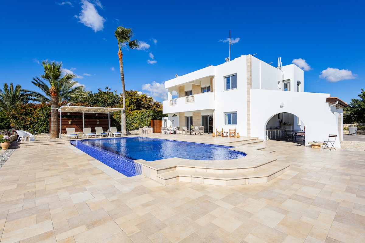 A modern villa exterior is shown, highlighting a large swimming pool with a unique shape and tiled edges. Surrounding the pool are sun loungers and a covered outdoor dining area, set against a backdrop of palm trees and a clear blue sky.