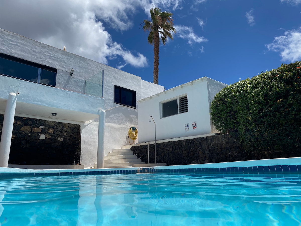 The image captures a private swimming pool with clear blue water reflecting the sky. Surrounding greenery and a palm tree are visible, along with a white building with large windows and stairs leading to the pool area. The scene suggests a tranquil outdoor space.