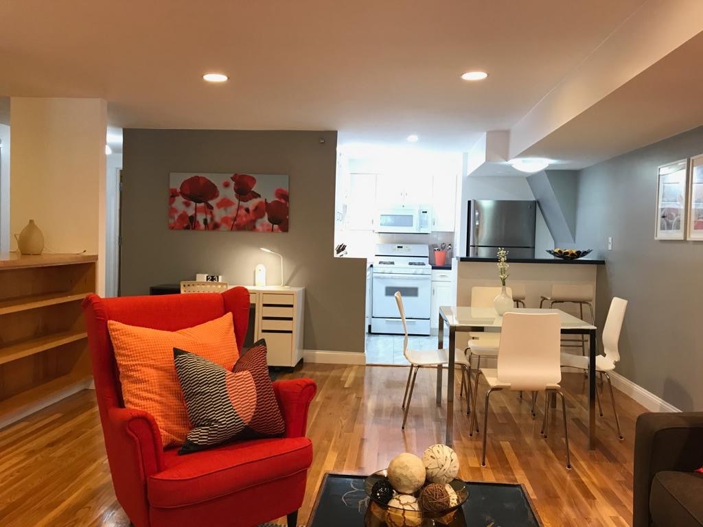A modern studio apartment is showcased with gray walls and warm wooden flooring. A bright red chair with patterned cushions is positioned near a dining table with white chairs. A full kitchen is visible, featuring stainless steel appliances and decorative items on display.