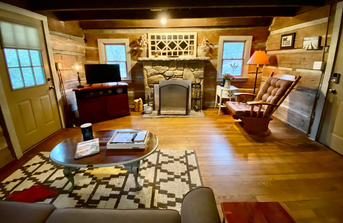 The living room features exposed log walls and warm wooden flooring. A stone fireplace serves as a focal point, surrounded by two windows. A rocking chair and television are positioned next to a coffee table, adding to the cozy atmosphere of the space.
