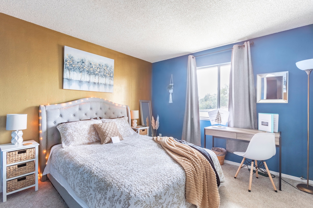 A spacious bedroom showcases a plush bed adorned with a neutral-colored quilt. Soft lighting accents the upholstered headboard, while a working desk stands by a window, allowing for natural light. Decorative elements include a blue and gold color scheme with thoughtful artwork on the walls.