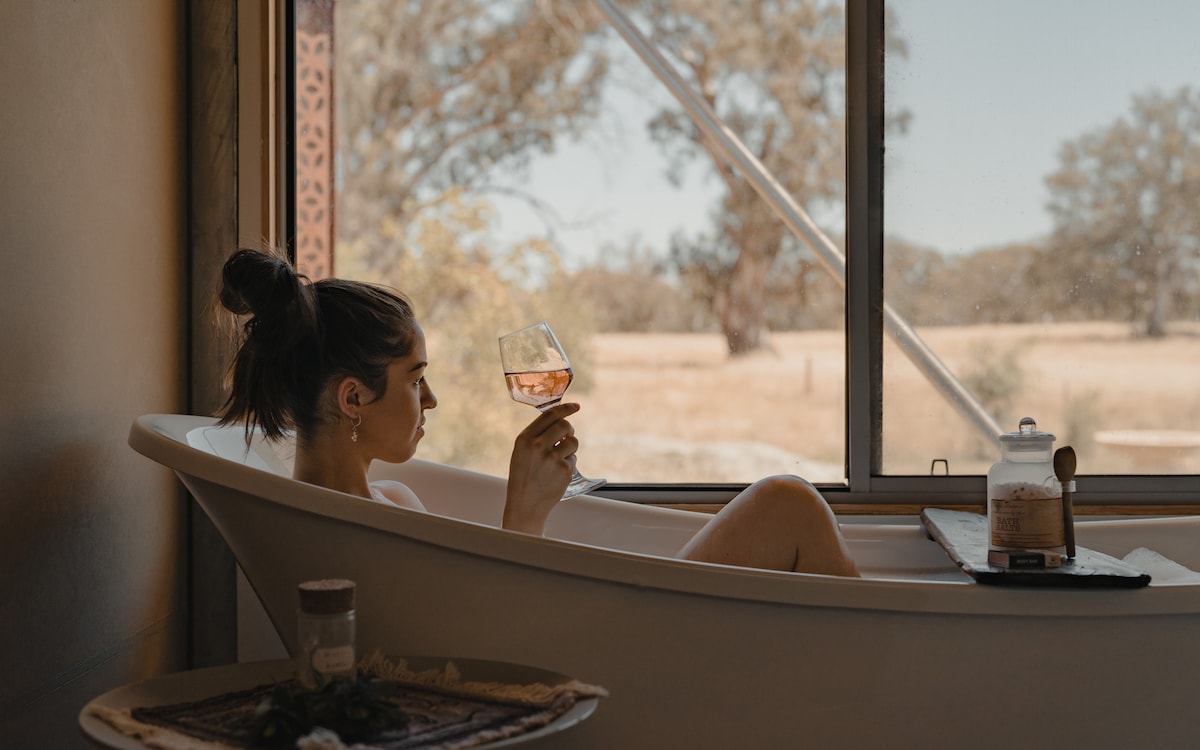 A claw foot bathtub is positioned beside a large window, revealing scenic views of the surrounding landscape. A glass of rosé is held, and a wooden tray nearby displays a candle and additional items, creating a serene ambiance for relaxation.