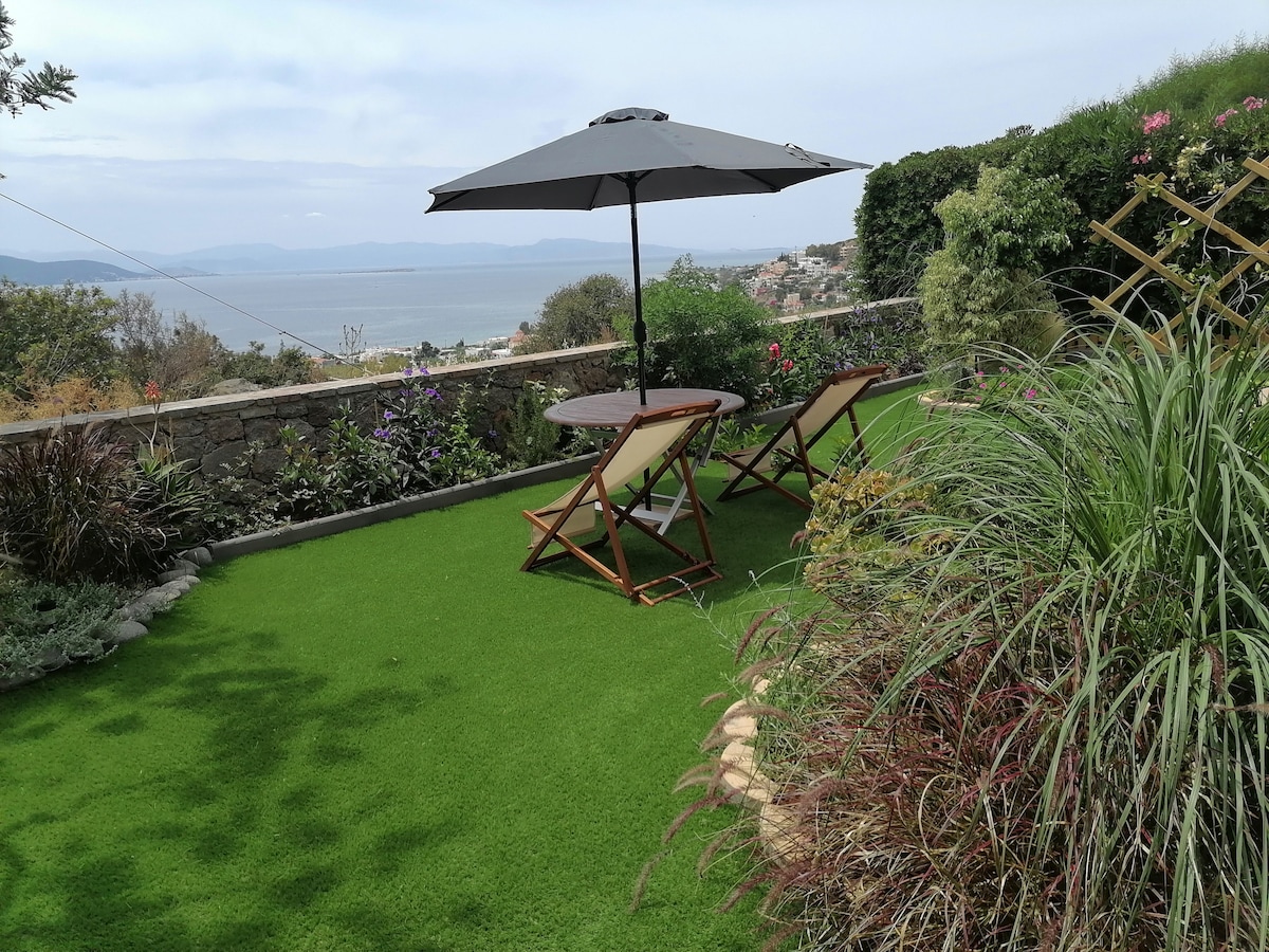 A well-maintained yard features a round table and two folding chairs beneath a large umbrella. Lush greenery and colorful flowers surround the area, with a scenic view of the Saronic Gulf visible in the background.