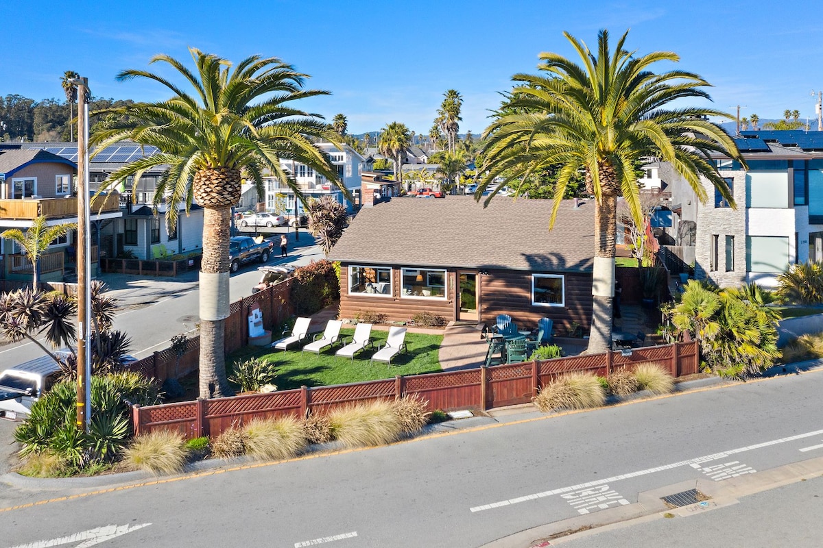 An aerial view of a charming bungalow is surrounded by lush landscaping and towering palm trees. The exterior features a welcoming front yard with lounge chairs positioned on green grass, framed by a wooden fence. Nearby streets and other residences provide a sense of neighborhood.