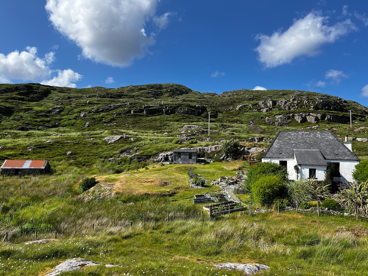 The image captures a serene landscape featuring lush green hills under a bright blue sky with fluffy white clouds. A charming cottage is prominently displayed, surrounded by a grassy area. Nearby structures, including a rustic shed with a red roof, complete the tranquil scene.