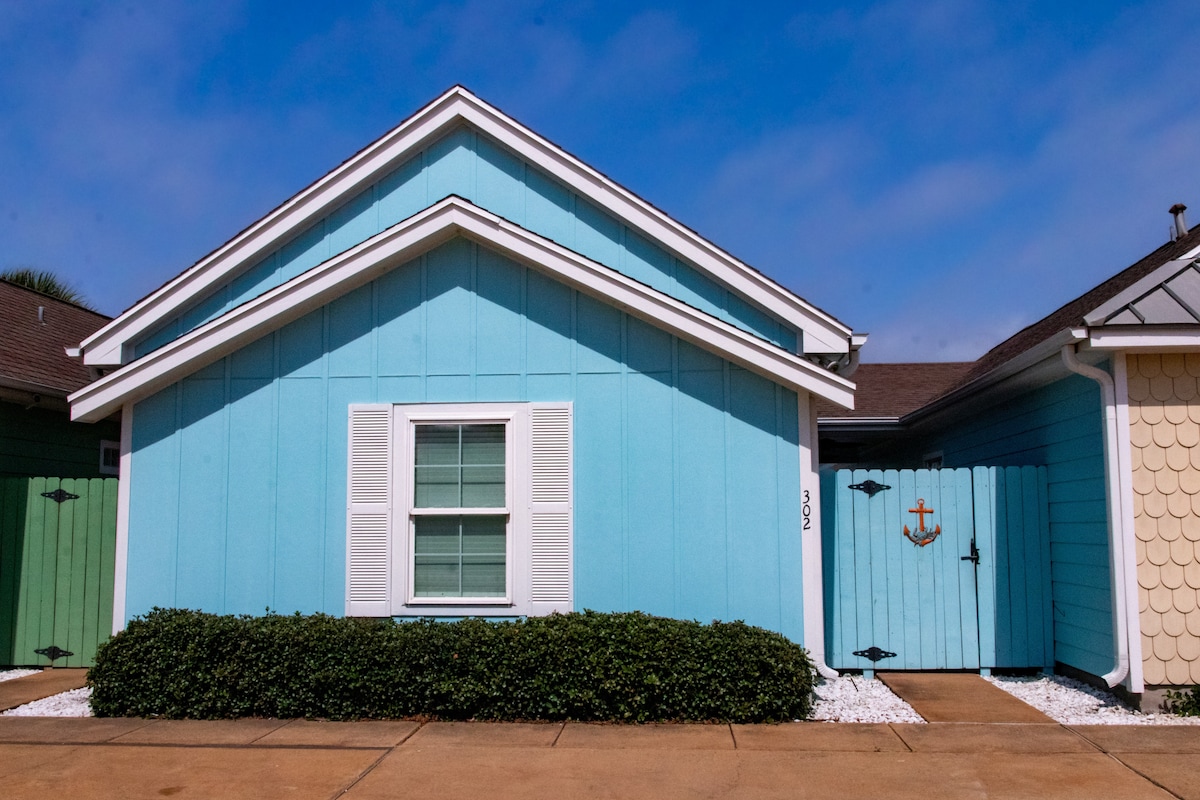 The exterior of a light blue townhome is depicted, featuring white shutters on a central window. A neatly trimmed green hedge borders the front, while a gated entrance with a minimal anchor design is visible to the right.
