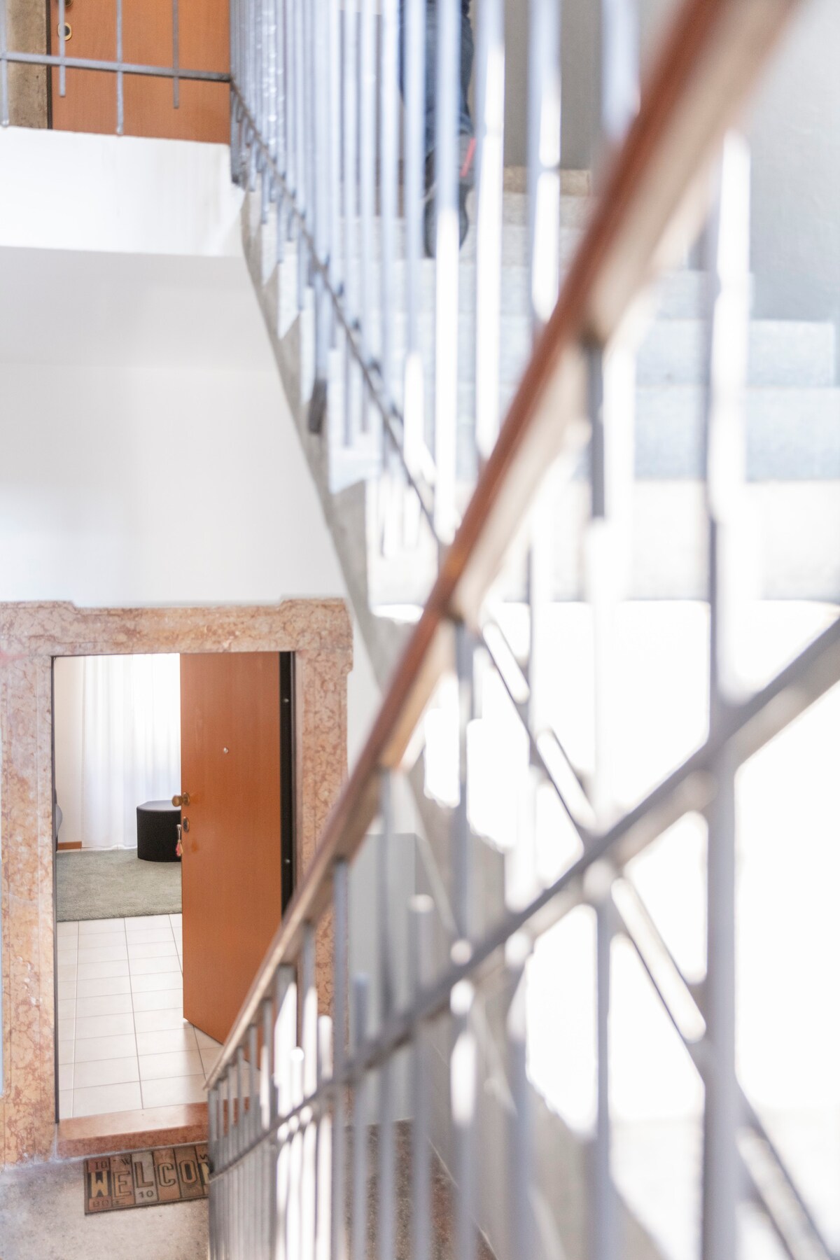 A well-lit staircase features a polished handrail and stone steps leading to the second floor. An inviting entrance is visible at the bottom, showcasing a wooden door with a subtle welcome mat positioned in front. Natural light illuminates the space, emphasizing its clean lines and modern design.