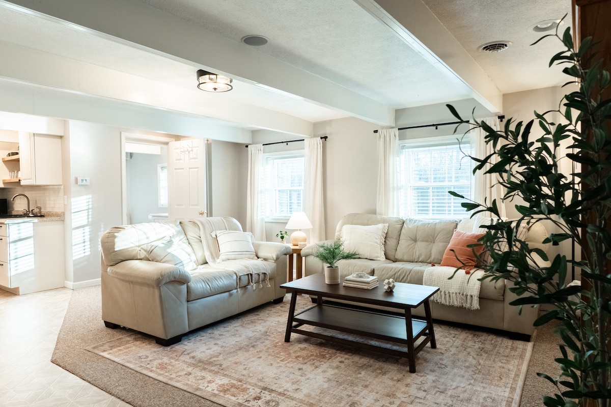 A spacious living room features cream-colored sofas and a wooden coffee table. Natural light filters through large windows, illuminating the soft rug underneath. A small plant and lamp add a touch of greenery and warmth, creating a comfortable space for relaxation.