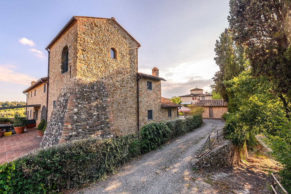 An ancient farmhouse stands proudly, showcasing its rustic stone façade and unique tower. A gravel path leads toward the inviting entrance, surrounded by lush greenery and terracotta-tiled areas. The landscape captures the serene charm of the Tuscan countryside, with rolling hills in the background.
