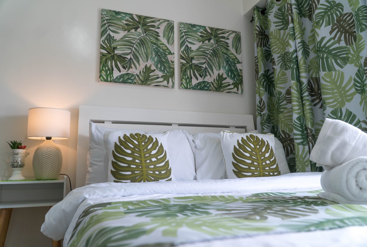 The bedroom is adorned with leafy wall art above a queen bed, which is dressed in white linens featuring green foliage patterns. Decorative cushions echo the room's tropical theme, complemented by a coordinating curtain. A bedside lamp provides soft lighting, enhancing the calm ambiance.