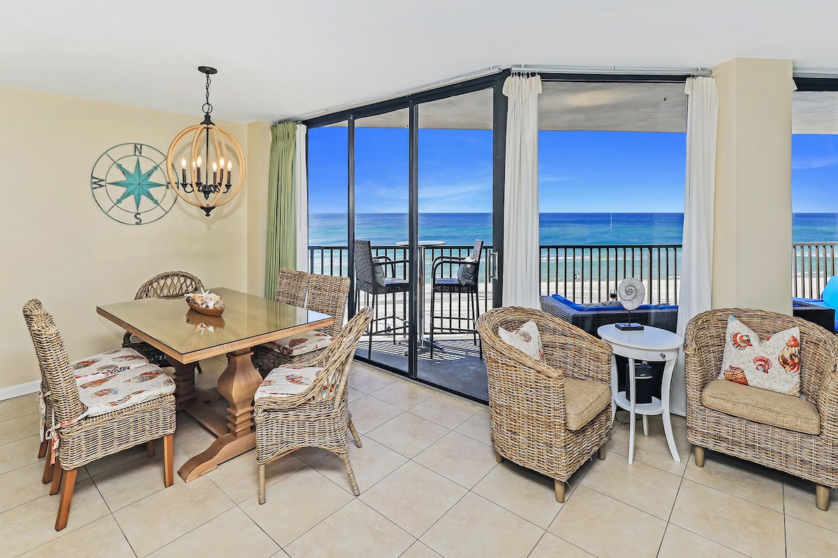 A dining area features a large wooden table surrounded by wicker chairs with cushions. Floor-to-ceiling windows reveal a stunning ocean view, allowing natural light to fill the space. Coastal décor is subtly integrated throughout the area, creating an inviting atmosphere.