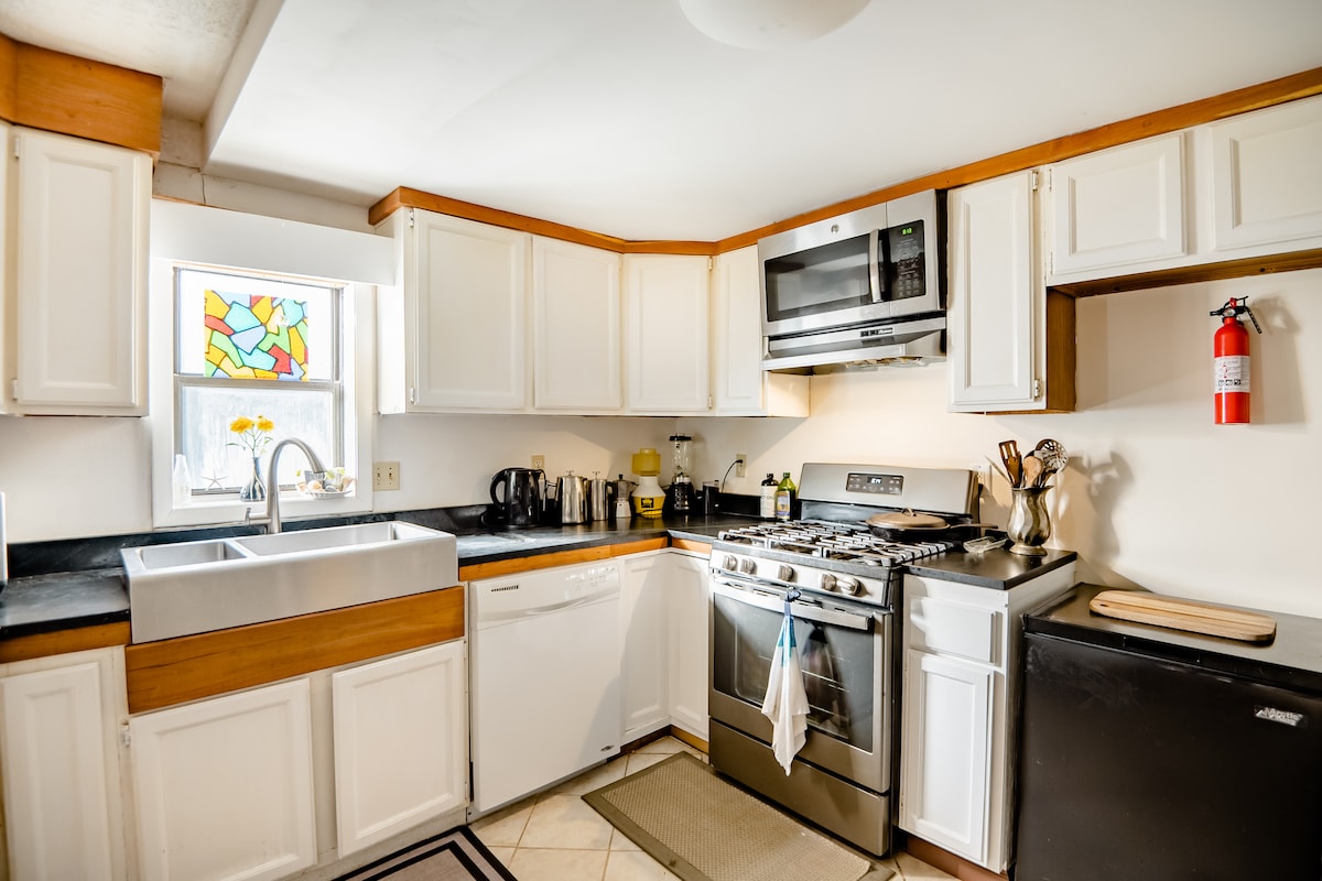 A well-equipped kitchen features ample cabinet space, a stainless steel gas stove, and a large sink beneath a stained glass window. Various kitchen utensils and a coffee maker are visible, contributing to a functional cooking environment.