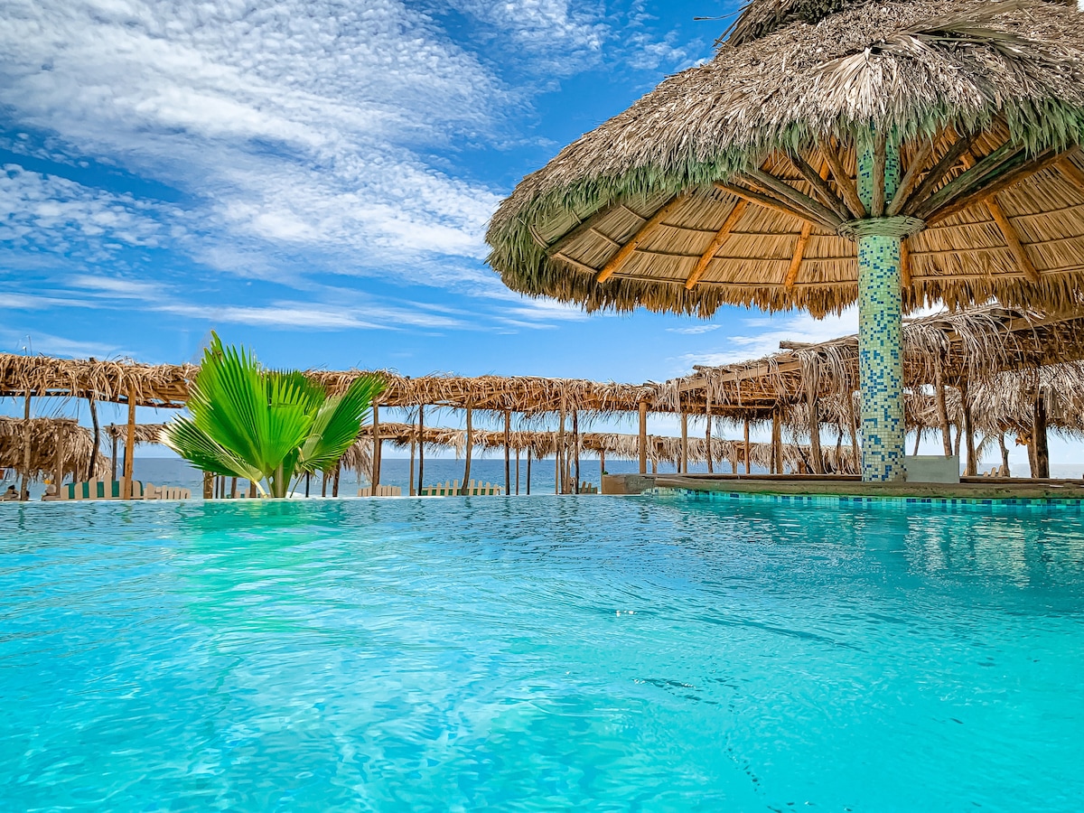 A refreshing pool area is surrounded by palm trees and thatched-roof structures. The tranquil water reflects the blue sky, while shaded seating areas provide a relaxing space to enjoy the view. Lush greenery enhances the inviting atmosphere of the outdoor space.